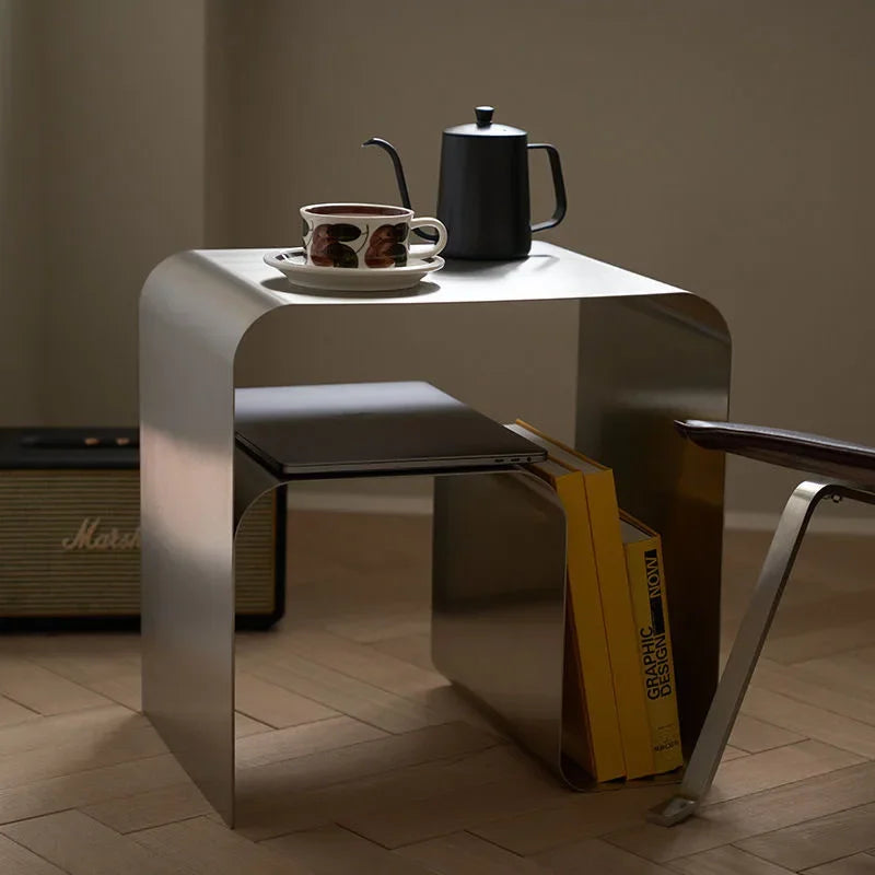 A brushed silver, bent metal side table with a black pour-over kettle and a coffee cup on top, and books stored below.