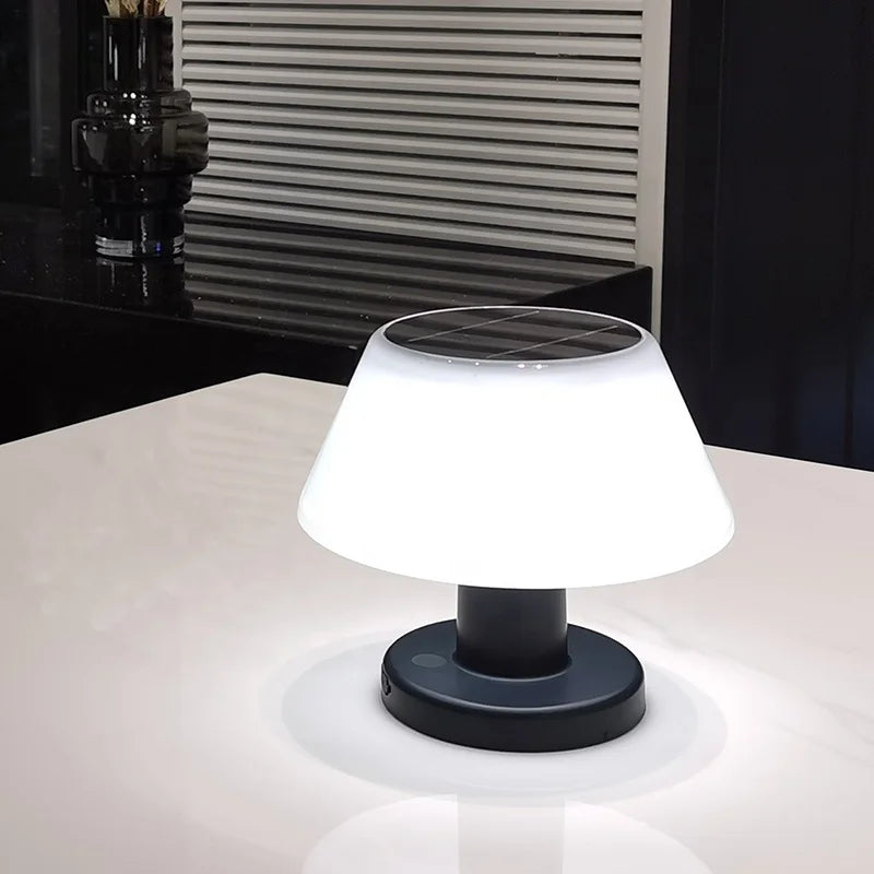 A small, solar-powered table lamp with a white, slightly tapered shade and a dark base illuminates a white surface. The lamp's top features a visible solar panel. The background is blurred but shows a dark surface with a decorative black object and a window with horizontal blinds.