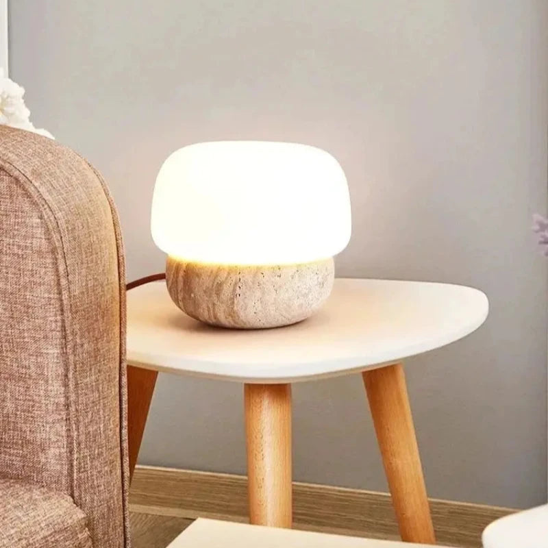 A small, glowing table lamp with a rounded white shade and a textured, light-colored base sits on a small white side table with light wooden legs. The lamp emits a warm light, and its brown cord extends to the left. The table is positioned next to a brown upholstered armchair. The background is a light grey wall.