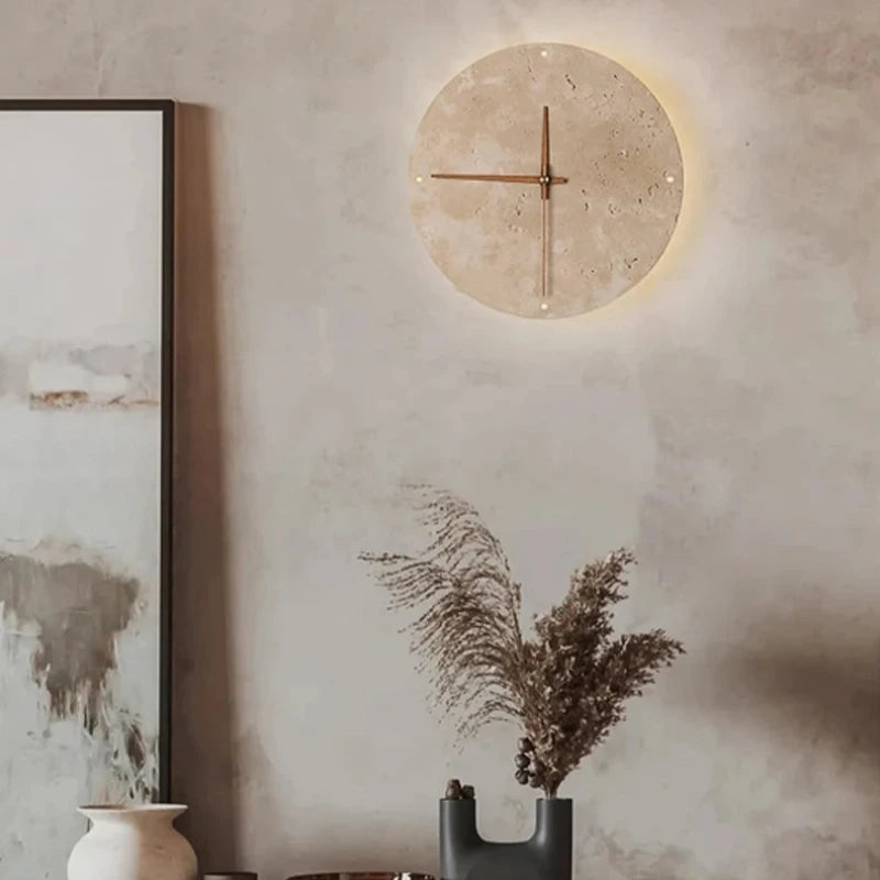 A round wall clock with a light-colored, textured face and simple brown hands is mounted on a textured grey wall. The clock is softly backlit, creating a subtle glow around its perimeter. Below the clock, a dark, uniquely shaped vase holds dried feathery grasses. Part of a framed abstract artwork is visible on the left.
