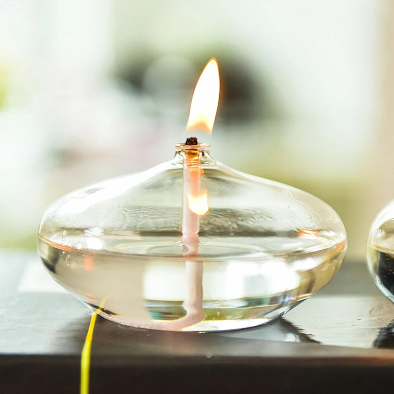 A small, clear glass oil lamp with a lit wick sits on a dark, reflective surface. The flame is bright, and the lamp is partially filled with clear oil. Another similar lamp is partially visible on the right. A blurred background suggests a bright, possibly outdoor setting.