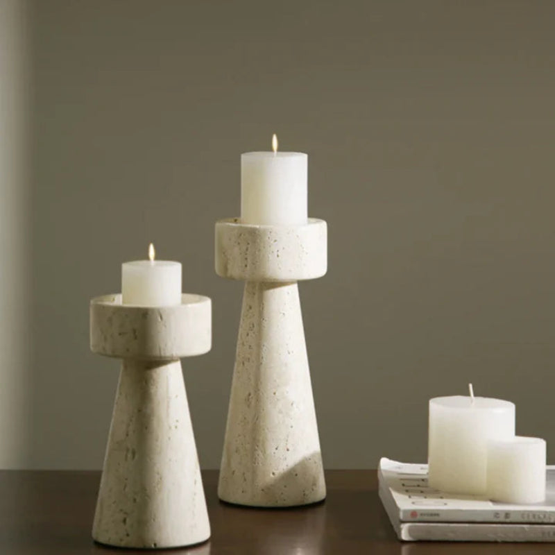 Two textured, light-colored faux travertine stone candle holders of varying heights, each holding a lit white pillar candle, arranged on a dark surface with two unlit white pillar candles resting on a stack of magazines.