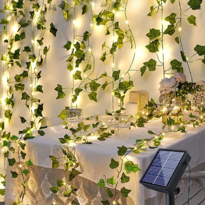 A curtain of warm white solar-powered LED lights intertwined with artificial green ivy leaves hangs on a wall behind a table covered with a white lace tablecloth. The lights also drape across the table, illuminating glasses and decorative items. A small solar panel is visible on the right.
