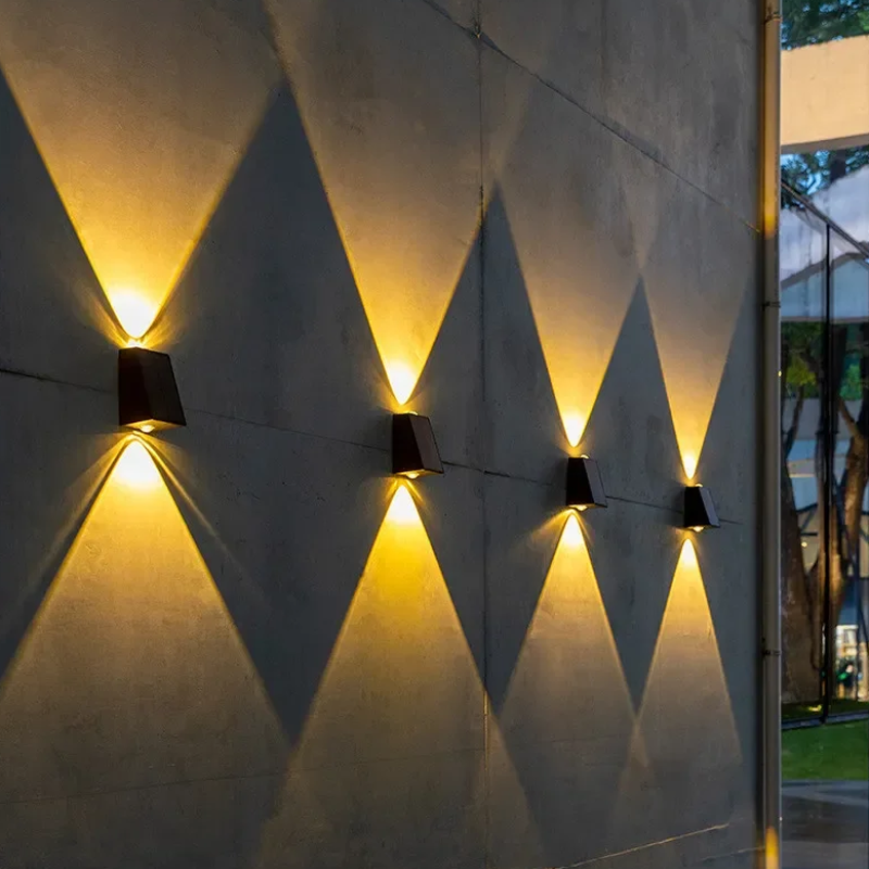 Four small, dark, rectangular lights are evenly spaced and mounted on a textured wall, each casting a warm white triangular beam both upwards and downwards at dusk or night. A reflecting pool runs along the base of the wall.