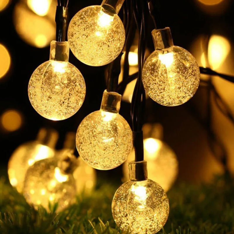 A close-up shows several round, textured string lights emitting a warm glow, hanging against a dark background with blurred bokeh lights and a hint of green foliage below.