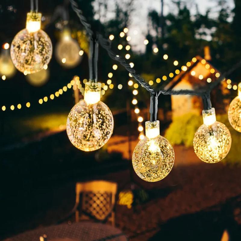 A close-up shows a string of glowing, textured round lights with a warm color temperature in an outdoor setting at night. Blurred strings of smaller lights create a bokeh effect in the background. The suggestion of a chair is visible below.