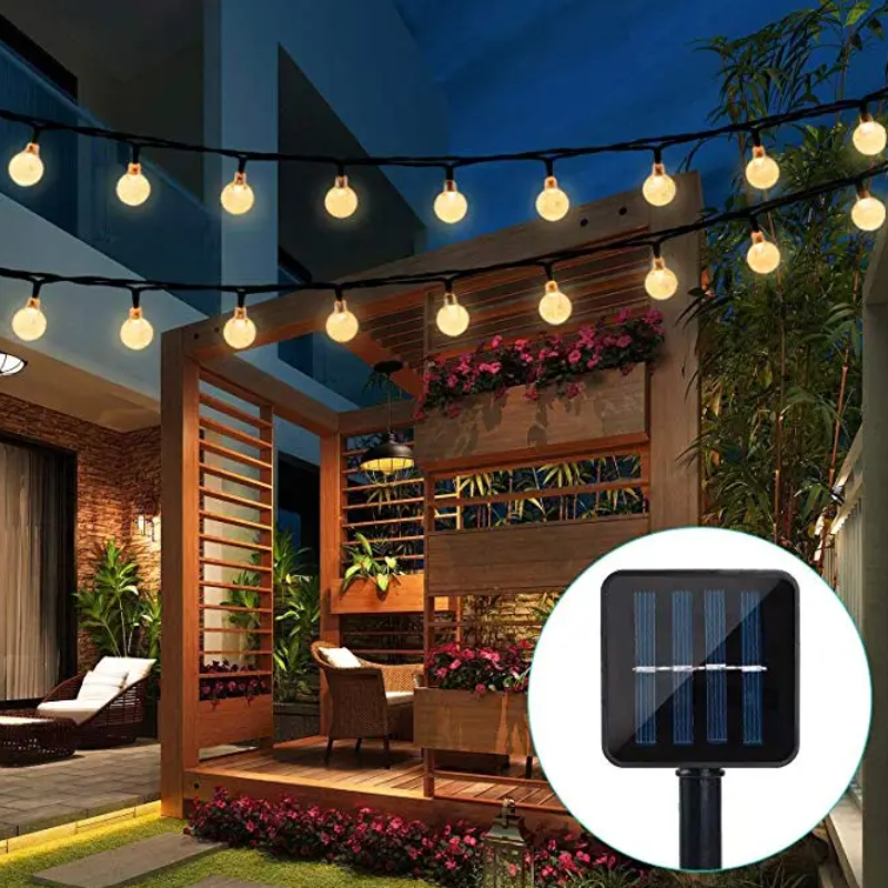 Two strings of round, warm-toned lights illuminate a patio area with wooden structures, plants, and outdoor furniture at dusk. A solar panel for the lights is shown in a circular inset.
