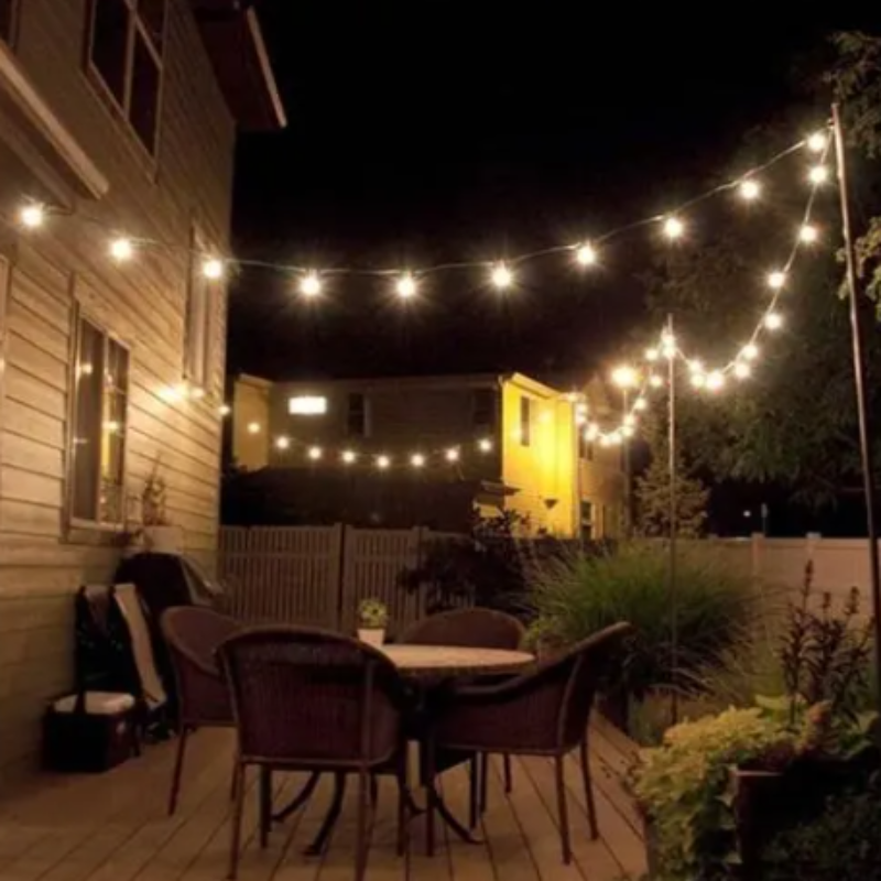 A string of glowing, warm-toned lights illuminates a backyard patio at night. A round table with chairs sits on a wooden deck next to a house. Other houses and trees are visible in the background.
