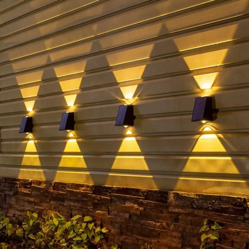 Four small, dark, rectangular lights are evenly spaced and mounted on a horizontally paneled wall above a brick base, each casting a warm white triangular beam both upwards and downwards at dusk or night. Green plants grow at the base of the wall.