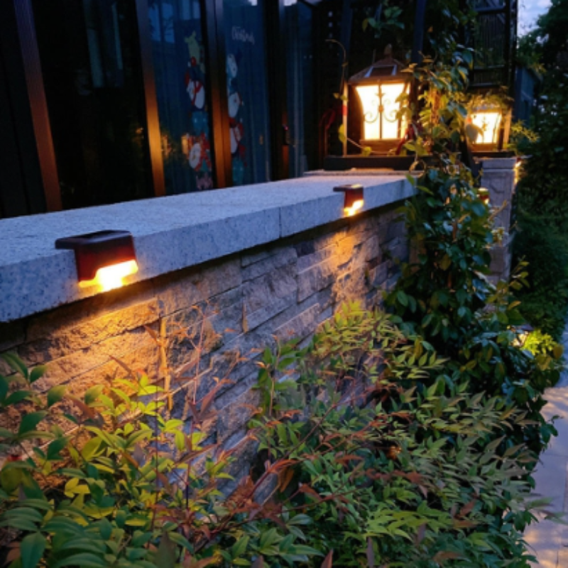 A stone garden wall at night is illuminated by two small, dark, semi-circular solar lights mounted along its top edge, casting a warm downward glow onto the textured stone. Lush greenery and a few taller lantern-style lights are visible behind the wall, enhancing the nighttime garden ambiance. The foliage in the foreground appears to be Nandina domestica, also known as heavenly bamboo.