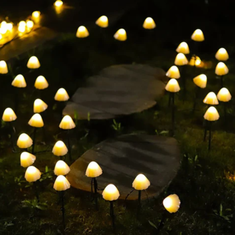 Numerous small, mushroom-shaped solar lights with translucent caps emit a warm yellow glow in a garden at night. The lights are on thin black stakes and are scattered among dark foliage and stepping stones. String lights with small bulbs are visible in the upper left corner.