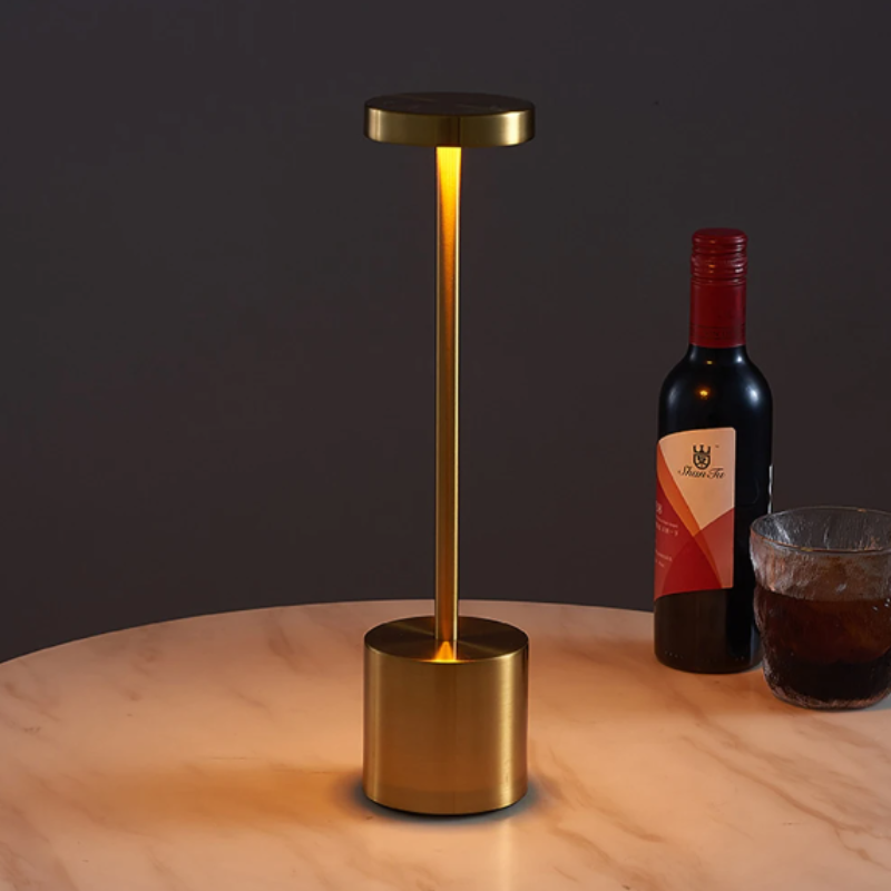A cordless LED table lamp with a brushed gold finish, featuring a cylindrical base, a slender stem, and a circular light panel emitting a warm, downward glow, situated on a round marble-patterned table next to a bottle of red wine and a glass of amber liquid. The background is dark.