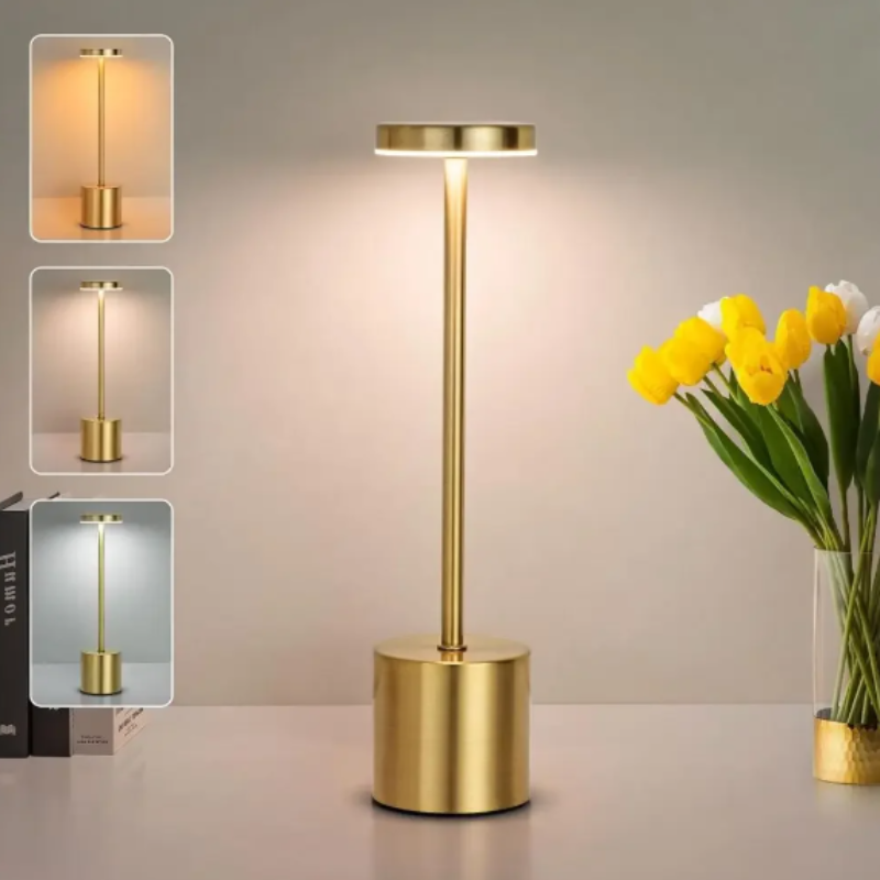 A sleek, cordless, rechargeable LED table lamp with a brushed gold finish, featuring a cylindrical base, a slender stem, and a circular light panel, emitting a warm glow. Three inset images show the lamp with different light color temperatures. The lamp is placed on a light-colored surface next to a vase of yellow and white tulips.