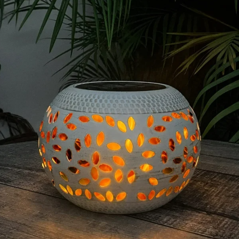 A round, off-white outdoor lantern with decorative oval cutouts emits a warm orange glow as it sits on a dark wooden surface. The lantern has a dark, flat top, likely a solar panel. Palm-like leaves are visible in the blurred background.