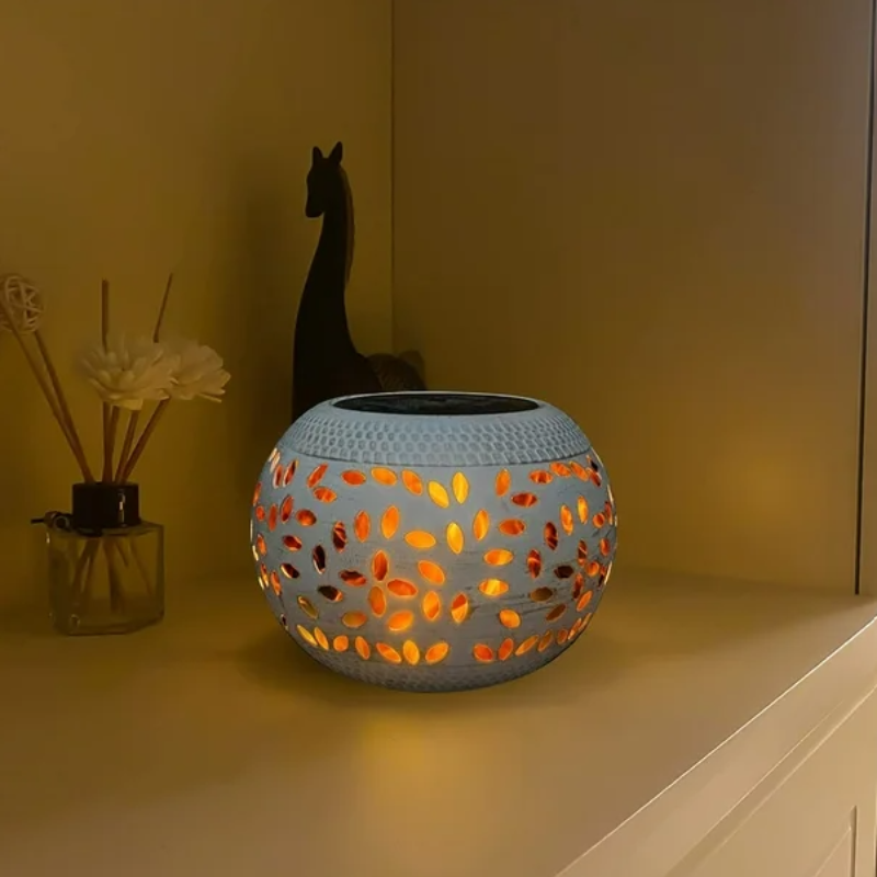 A round, off-white outdoor lantern with decorative oval cutouts emits a warm orange glow as it sits on a light-colored surface indoors. The lantern has a dark, flat top, likely a solar panel. A diffuser with sticks and a dark decorative object are in the blurred background.