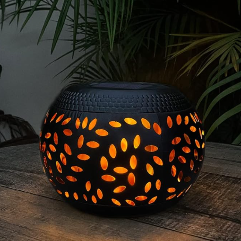 A round, black outdoor lantern with decorative oval cutouts emits a warm orange glow as it sits on a dark wooden surface. The lantern has a dark, flat top, likely a solar panel. Palm-like leaves are visible in the blurred background.
