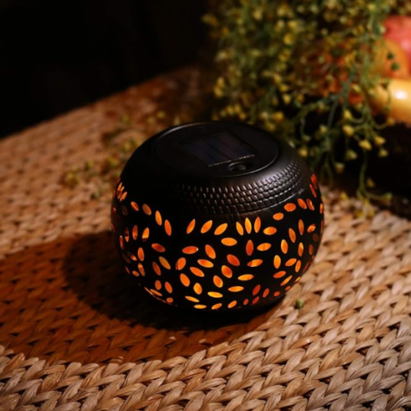 A small, round, black outdoor lantern with decorative oval cutouts emits a warm orange flickering light as it sits on a woven surface. The lantern has a dark, flat top, likely a solar panel. Blurred greenery is visible in the background.