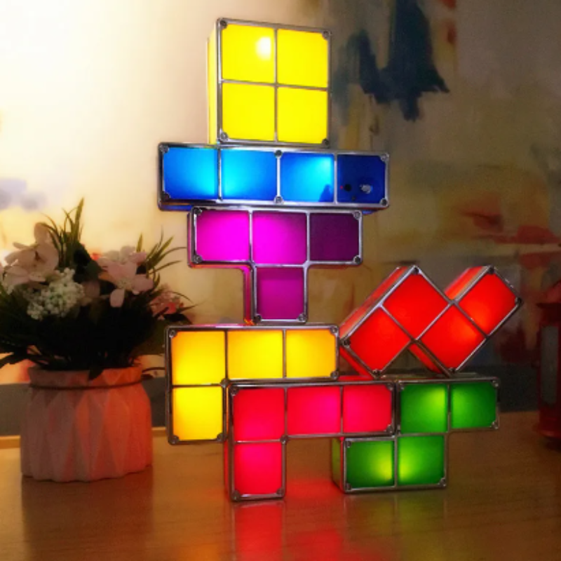 A stack of illuminated, colorful geometric block lights, reminiscent of a puzzle game, rests on a surface. The lights are in various shapes and colors including yellow, blue, purple, red, and green, creating a vibrant display. A potted plant with light-colored flowers is visible to the left of the light stack.