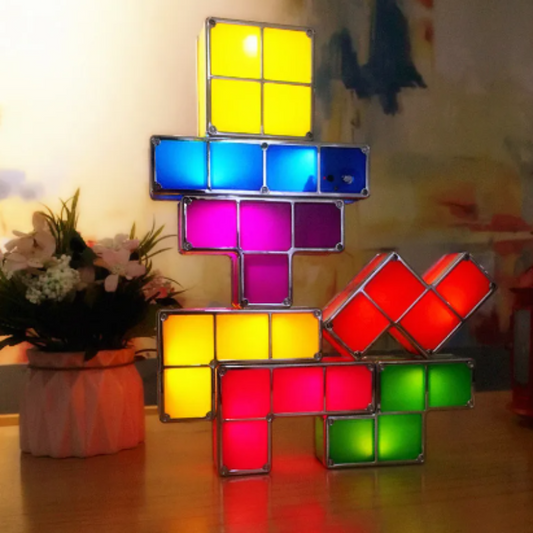 A stack of illuminated, colorful geometric block lights, reminiscent of a puzzle game, rests on a surface. The lights are in various shapes and colors including yellow, blue, purple, red, and green, creating a vibrant display. A potted plant with light-colored flowers is visible to the left of the light stack.