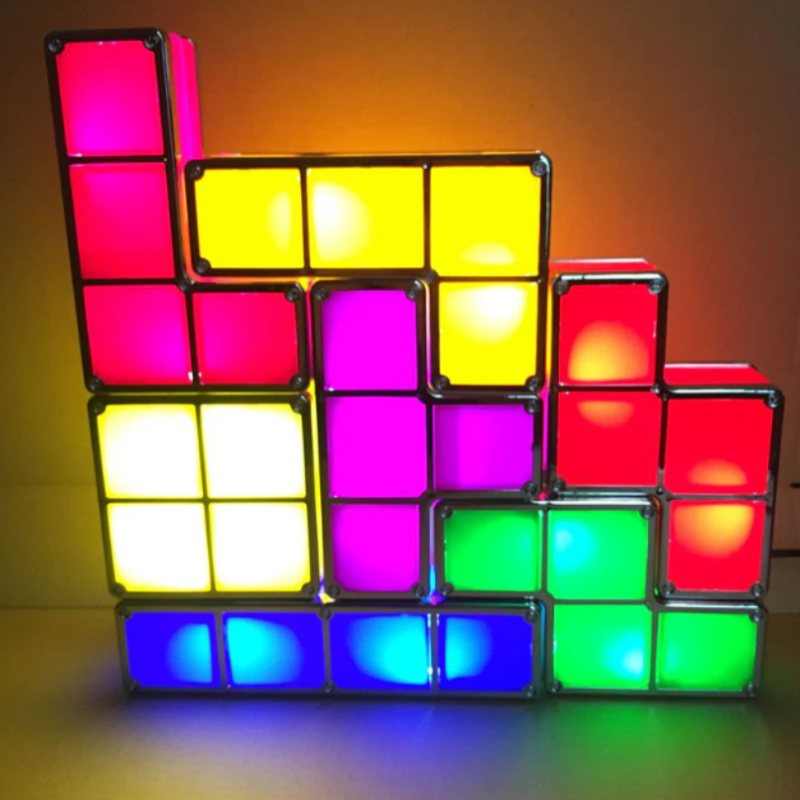 A colorful, illuminated structure made of interconnected geometric block lights, stacked in a staggered, stair-like arrangement. The blocks emit light in shades of pink, yellow, white, purple, blue, and green. They are placed on a surface, casting a soft glow beneath them.