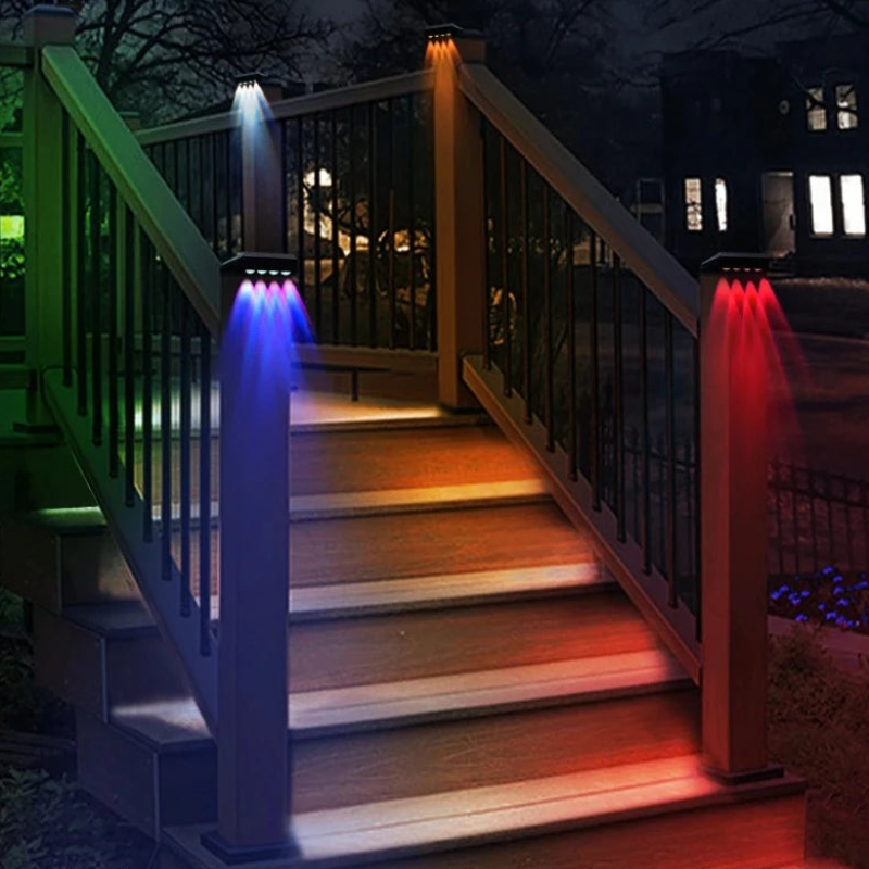 Four solar-powered post lights with color-changing LEDs illuminate a wooden staircase with railings at night. The lights emit white, blue, orange, and red beams downwards onto the steps and posts. A house with illuminated windows is visible in the background.