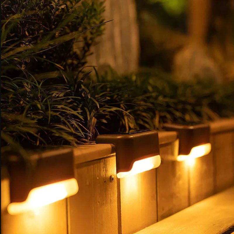 An outdoor staircase at night is illuminated by three small, dark, rectangular solar lights mounted on the sides of the steps. The lights cast a warm white glow downwards, lighting up the steps. The steps are made of wood and lead up to a house.