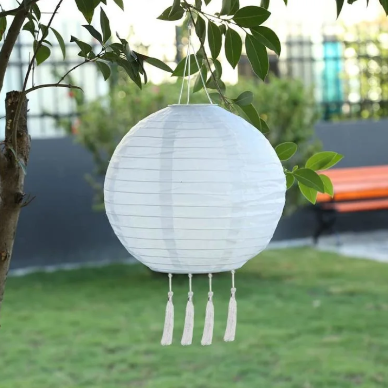 A round, white solar hanging lantern with tassels is suspended from a tree branch in an outdoor setting.