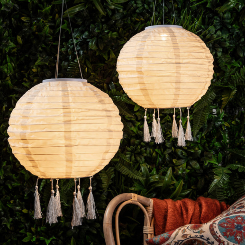The image shows two round, cream-colored paper lanterns hanging against a backdrop of green foliage. Each lantern has a series of tassels hanging from the bottom.