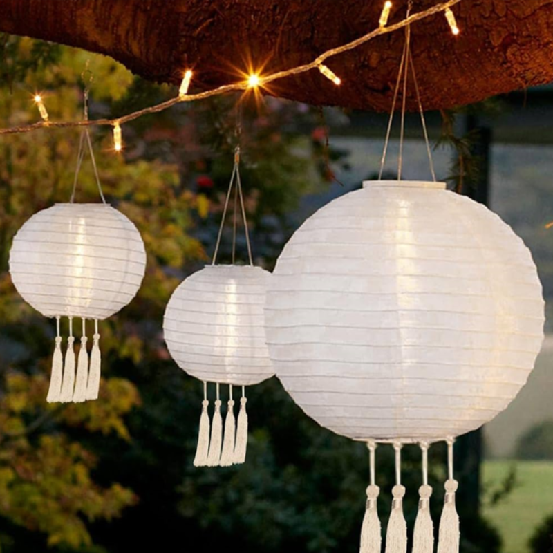 Three round, white paper lanterns with tassels hang from a tree branch, illuminated by string lights.