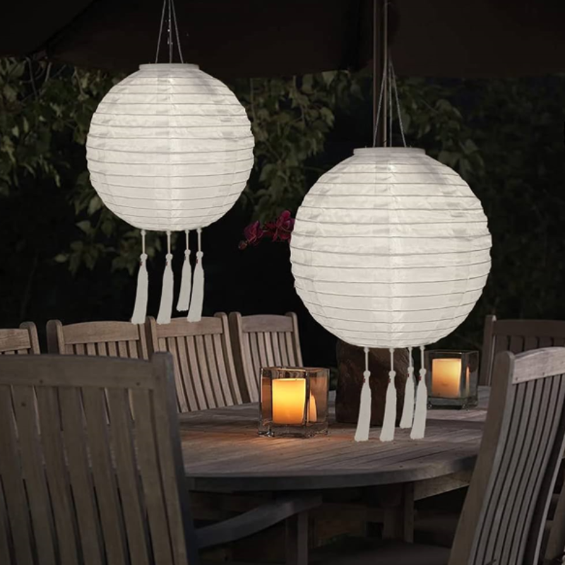 Two round, white solar lanterns with tassels hang above a wooden outdoor dining set with lit candles on the table at dusk.
