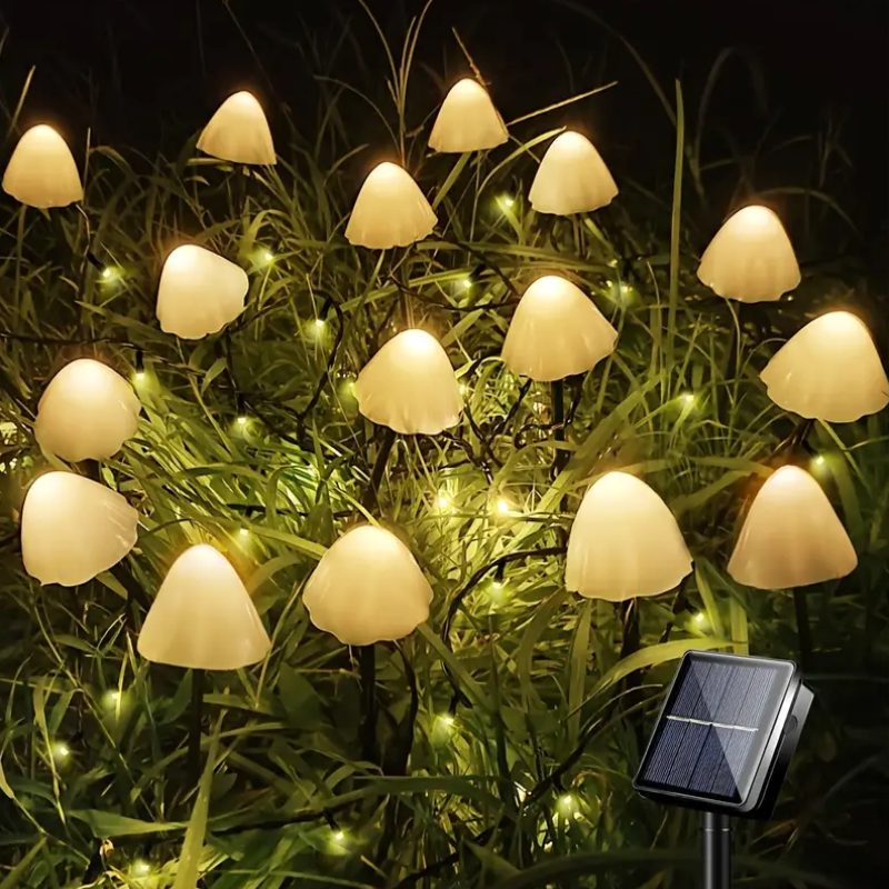 Numerous small, mushroom-shaped solar lights with translucent caps emit a warm white glow among green grass at night. A small square solar panel on a stake is visible in the lower right corner.