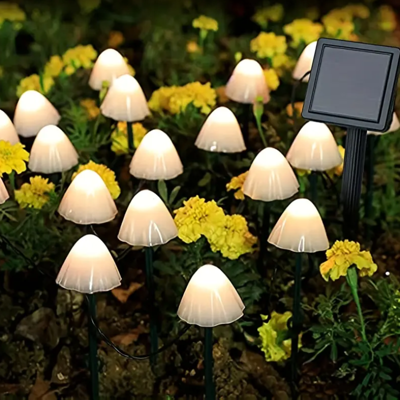 Numerous small, mushroom-shaped solar lights with translucent white caps emit a warm white glow among dark foliage and small yellow flowers at night. A small square solar panel on a stake is visible among the lights.