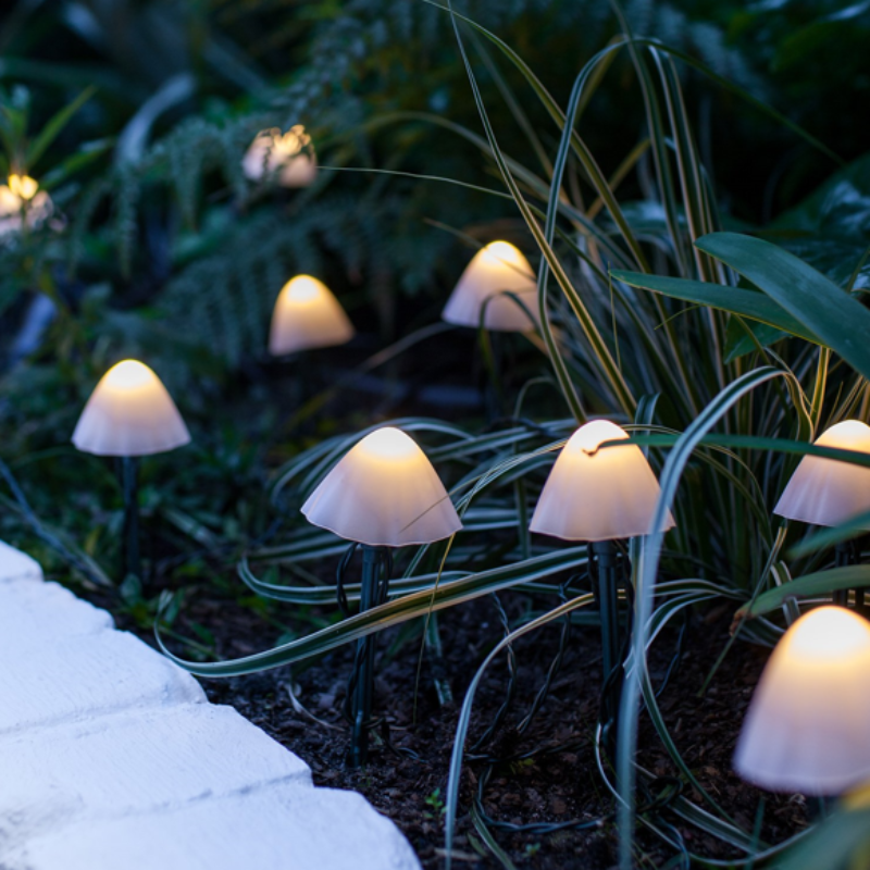 Several small, mushroom-shaped solar lights with translucent caps emit a warm white glow in a garden at dusk or night. The lights are on thin stakes and are scattered among dark foliage and along a light-colored stone pathway.