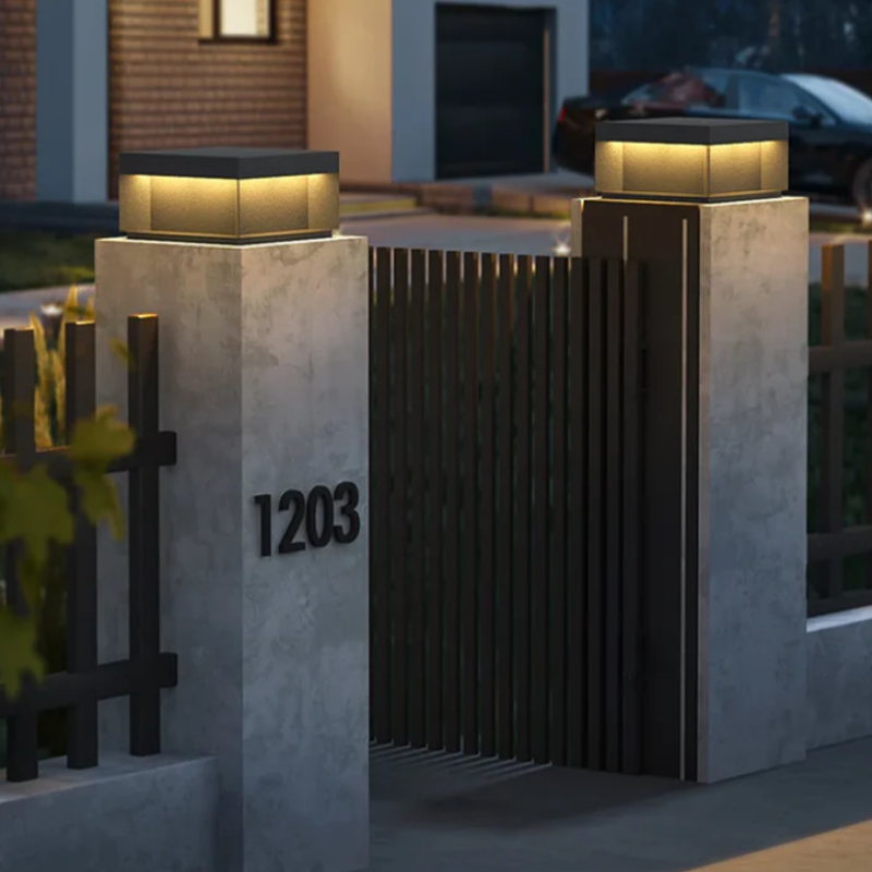 A modern gate entrance at dusk featuring gray concrete pillars with illuminated square lanterns on top and the house number "1203" displayed on one pillar.