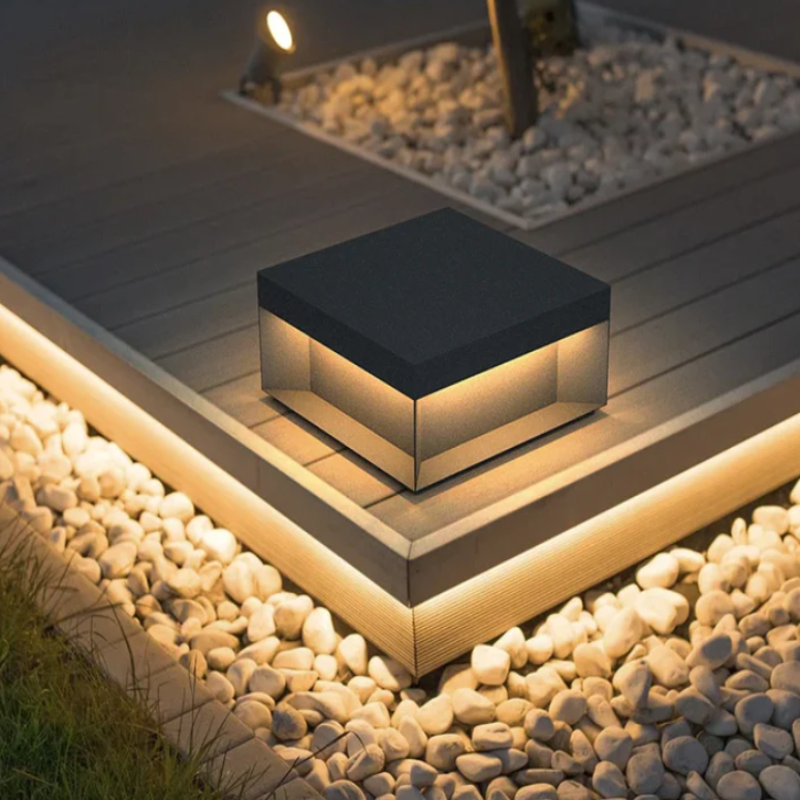 A contemporary, square outdoor light with a dark top and glowing sides, placed on a dark composite deck surrounded by white pebbles and soft underlighting.