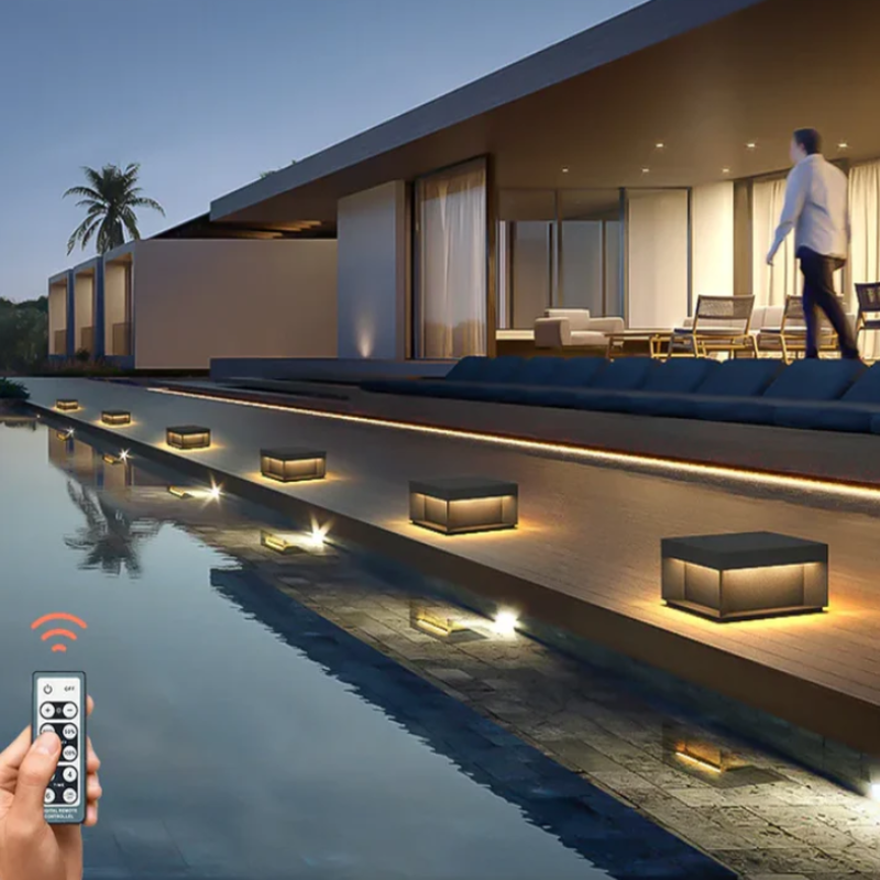A hand holding a remote control near a modern outdoor space at dusk, featuring a row of square lights with dark tops and glowing panels along a pool and deck, with a person walking on the illuminated patio.