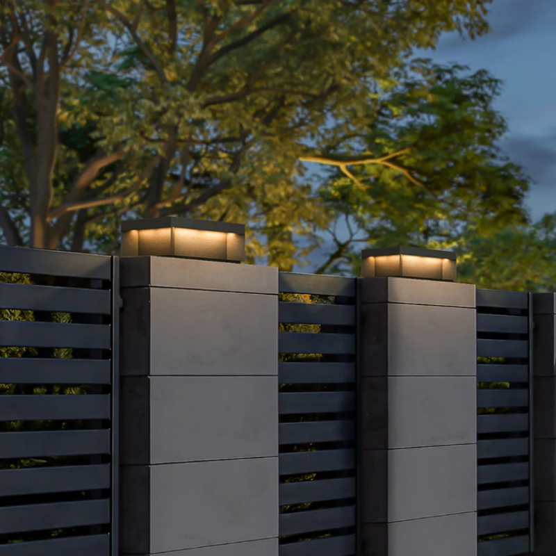 Two modern, square outdoor lights with dark tops and glowing warm-toned panels, mounted on top of gray fence posts at dusk.
