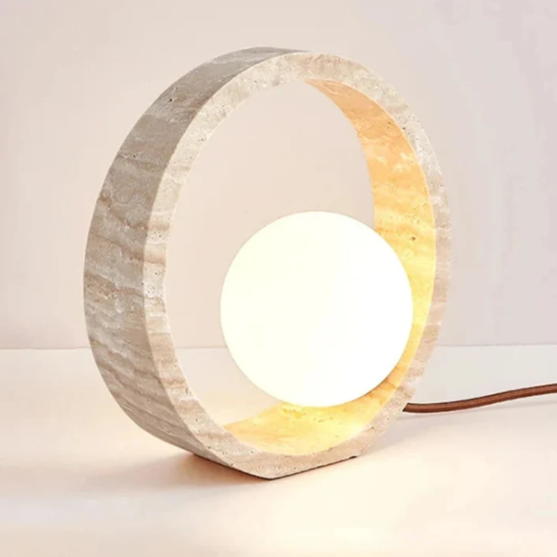 A circular table lamp features a textured, light-colored outer ring made of stone or concrete, enclosing a glowing white spherical light. The lamp sits on a light surface, and a brown cord extends to the right. The inner part of the outer ring is illuminated with a warm light.