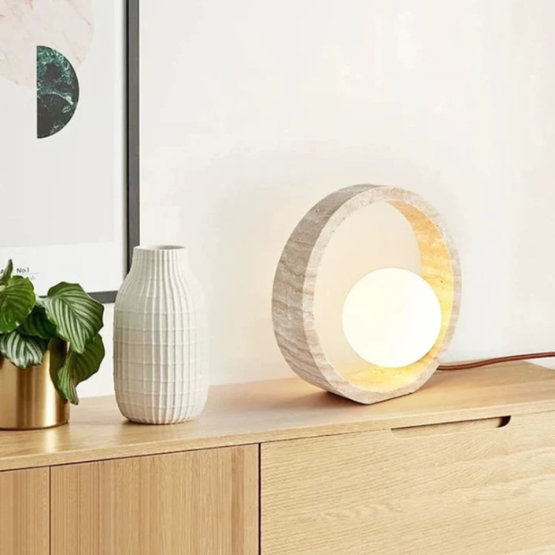 A circular table lamp with a textured, light-colored outer ring and a glowing white spherical inner light sits on a light wooden dresser. To the left of the lamp are a small gold pot with a green plant and a textured white vase. A framed piece of art is partially visible leaning against the wall behind the plant. The lamp's brown cord extends to the right against a white wall.
