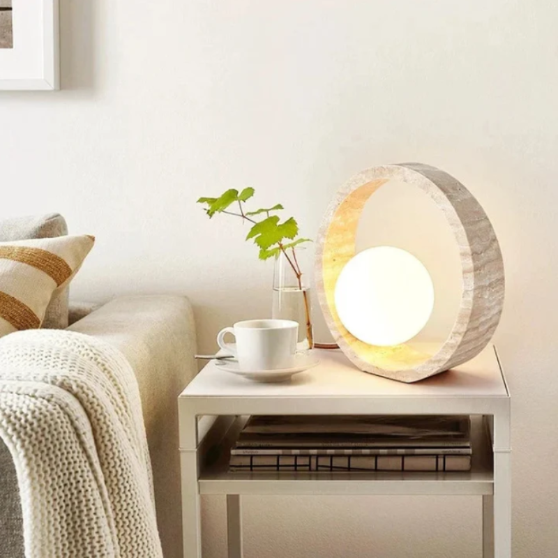 A circular table lamp with a textured, light-colored outer ring and a glowing white spherical inner light sits on a white nightstand. A white teacup on a saucer and a small clear vase with green leaves are also on the nightstand. Below, two books are stacked on a shelf. The nightstand is next to a bed with a patterned pillow and a light-colored knit blanket, against a white wall.