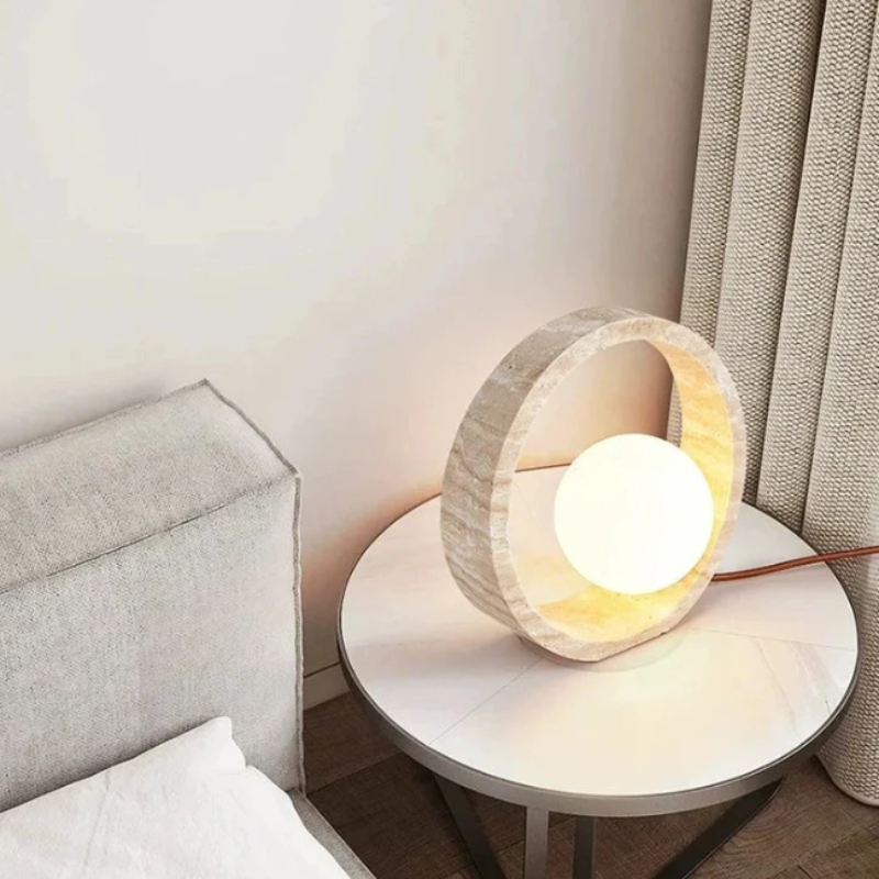 A circular table lamp with a textured, light-colored outer ring and a glowing white spherical inner light sits on a round side table with a light marble top and dark legs. The lamp's brown cord extends to the right. The table is positioned next to a bed with a light grey headboard and white linens, near a beige curtain. The inner part of the outer ring emits a warm glow.