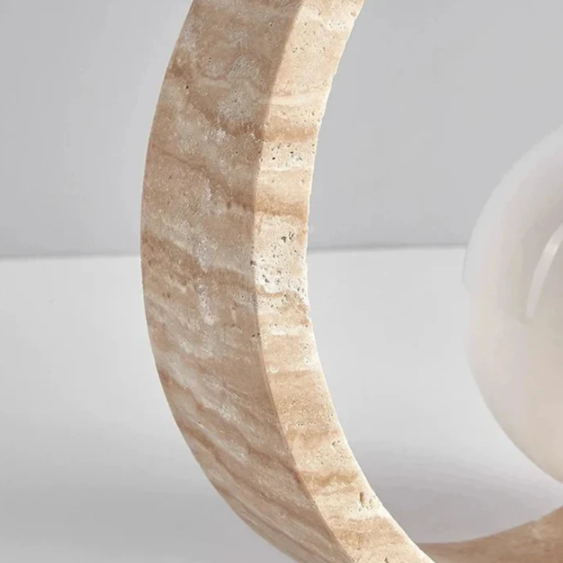 A close-up shows the textured, light-colored stone outer ring of a circular lamp, partially framing a glowing white spherical light source. The stone has natural variations in color and texture. A soft light emanates from behind the stone ring.