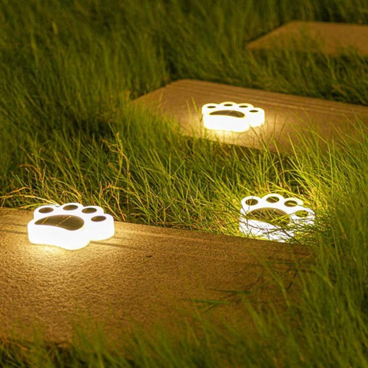Three small, white paw-shaped solar lights illuminate a stone pathway through grass at night, emitting a warm yellow glow from the paw pads and toes. The top of each light features a dark solar panel.