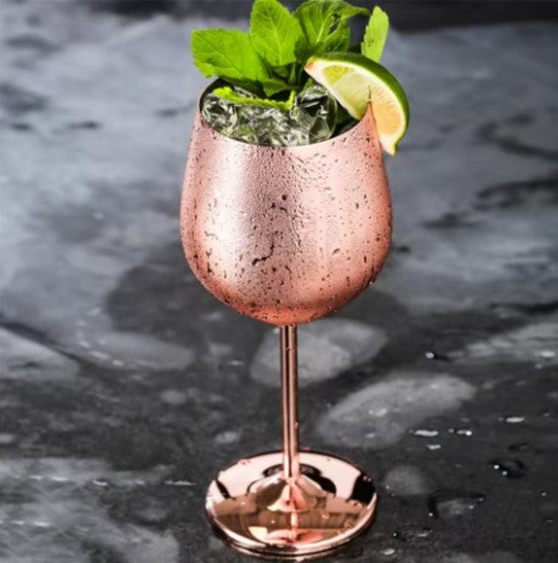 A single, elegant copper-toned stemmed glass is filled with ice, fresh green mint leaves, and a wedge of lime. The exterior of the glass has condensation droplets. It sits on a dark, textured surface that appears wet.