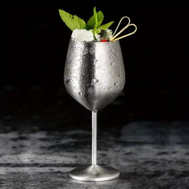A single, stemmed stainless steel cocktail glass is filled with ice, green mint leaves, and a clear liquid, possibly a cocktail. The exterior of the glass is covered in condensation droplets. A decorative pick with a heart-shaped end rests on the rim. The glass sits on a dark, textured surface against a blurred black background.