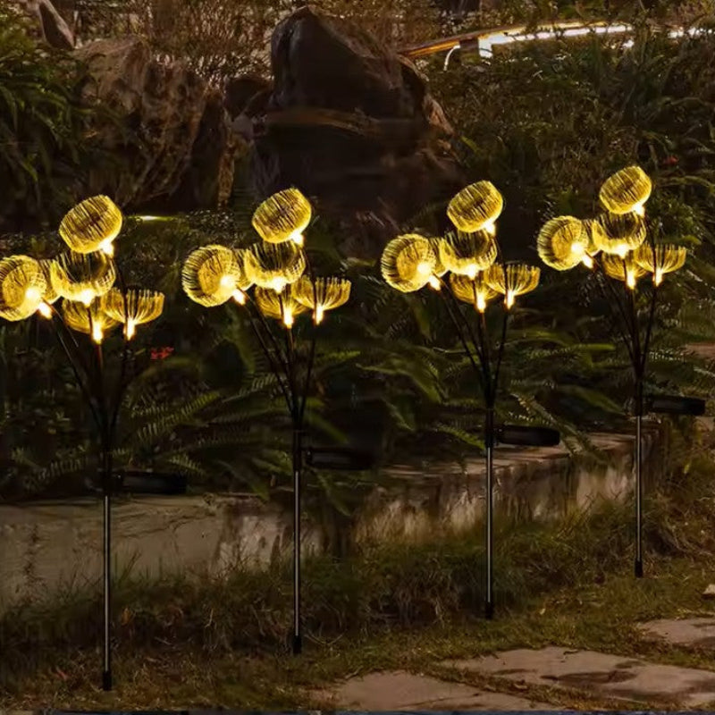 Four solar-powered garden light clusters with a glowing, jellyfish-like design emit a warm yellow light in a garden at night. They are on black stems and positioned near rocks and foliage.