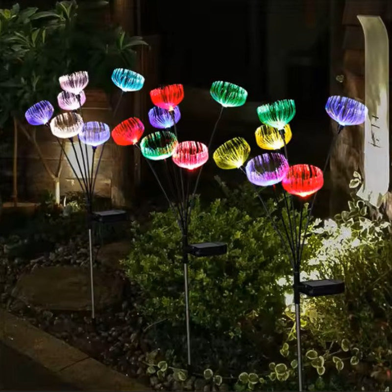 Several solar-powered garden lights with a jellyfish-like design emit a vibrant array of colors including purple, blue, green, yellow, and red in a garden at night. Each light cluster has multiple glowing tops on thin stems with a black solar panel base. They are situated among low-lying plants.