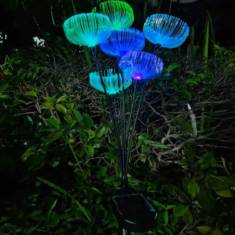 A cluster of solar-powered garden lights with a jellyfish-like appearance emits vibrant blue and green glows in a garden at night, rising from dark foliage. A black solar panel base is visible at the bottom.
