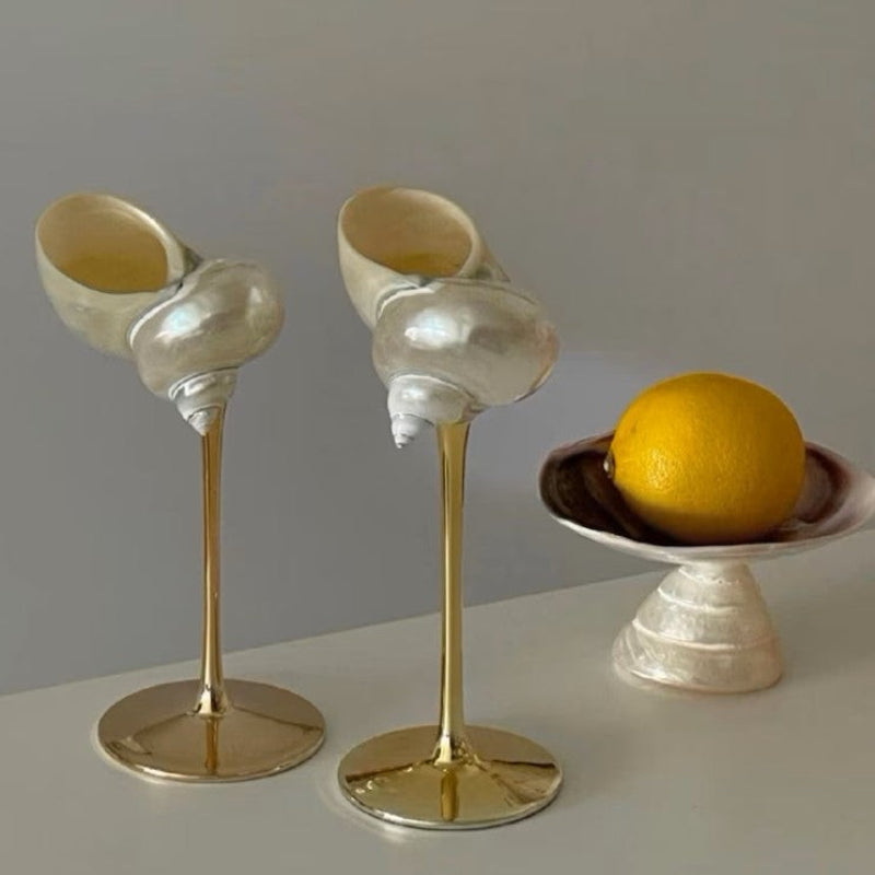 Two seashell wine glasses with gold stems and a small, decorative bowl made of seashell with a yellow lemon inside, all placed on a light-colored surface against a neutral background.