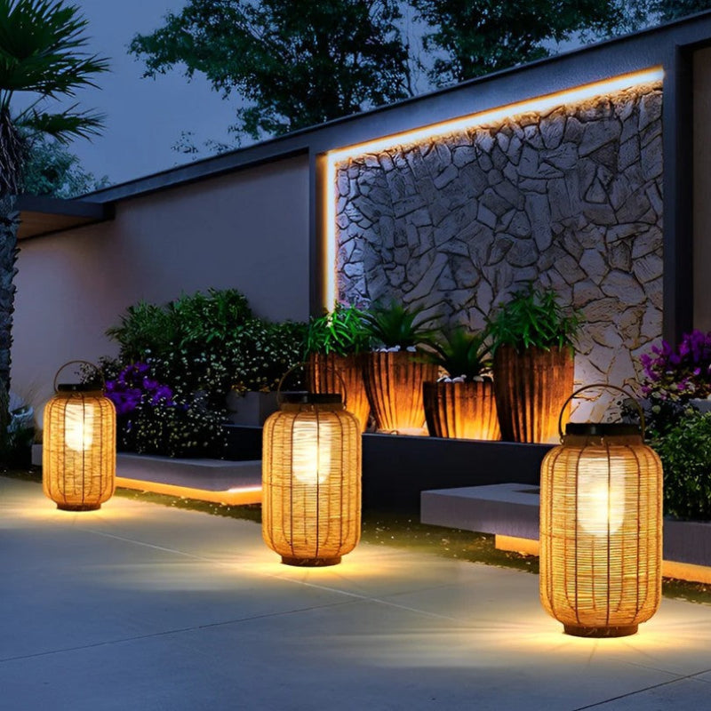 Three warm-toned, cylindrical outdoor lanterns made of woven material emit a soft white glow in a garden at dusk. The lanterns have dark tops, likely solar panels, and metal handles. They illuminate a paved area near a stone wall with uplighting, potted plants, and greenery.